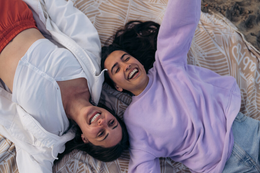view from two beautiful happy carefree young female friends tourists lying picnic blanket sandy beach smiling having fun enjoying vacation chilling outdoors looking relaxed